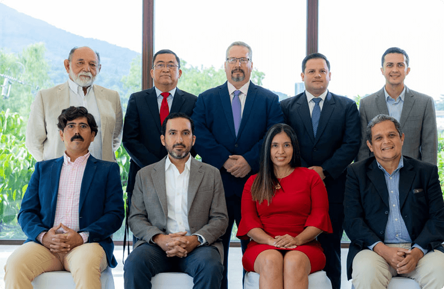 Diez personas, vestidas con traje de negocios, posan para una foto de grupo en un interior con grandes ventanales y vegetación visible al fondo.